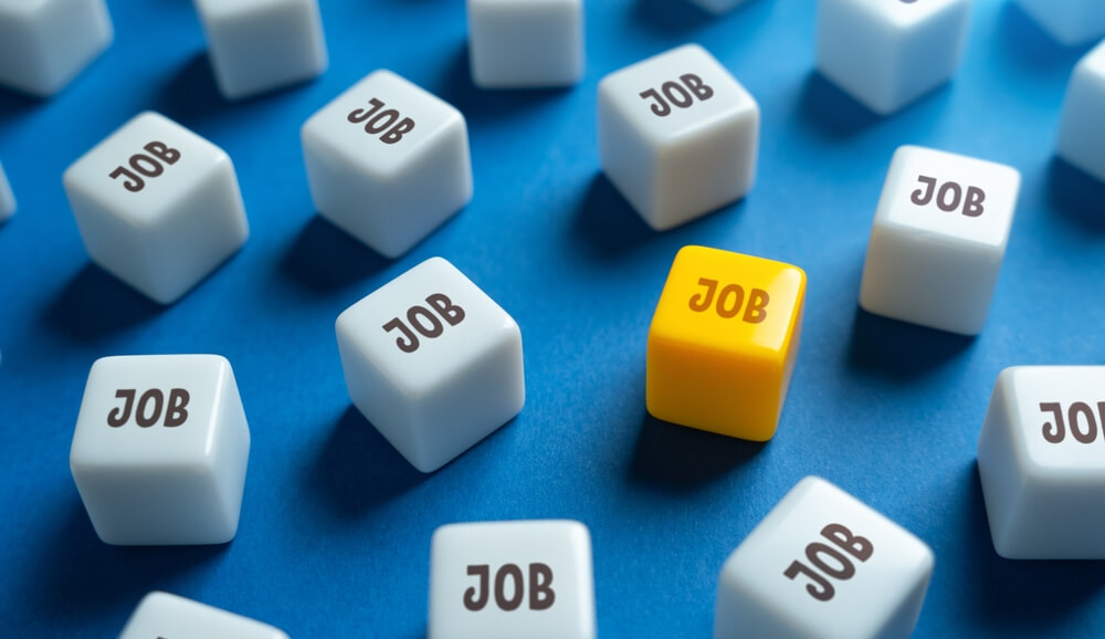 Multiple white dice with the word "JOB" in black are scattered on a blue surface. One yellow die, symbolizing a government position, stands out prominently among them.